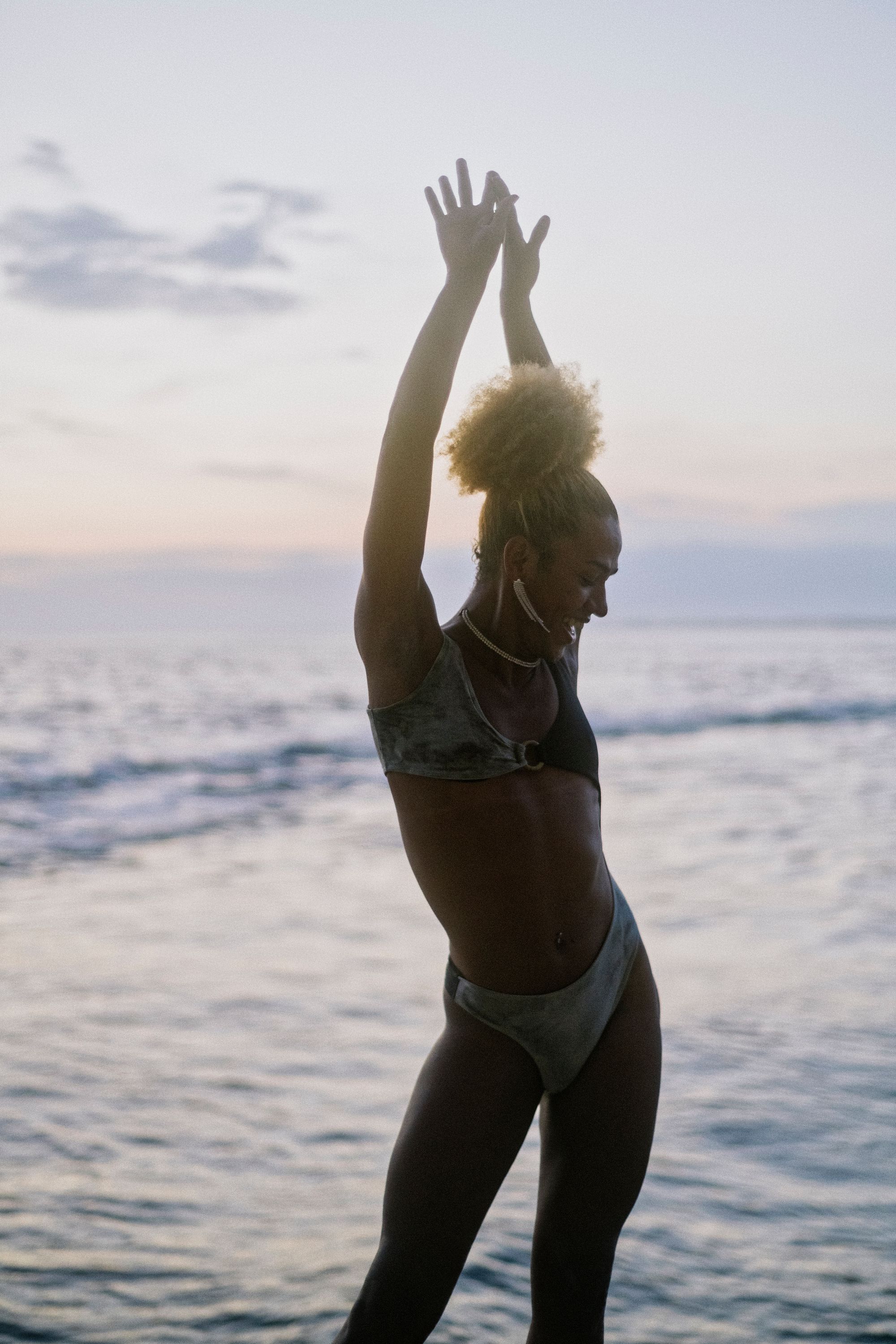 Brasilian travesti with natural red hair, wearing a crystal necklace, two piece bathing suit, poses in front of camera with hands raised up, with ocean in background