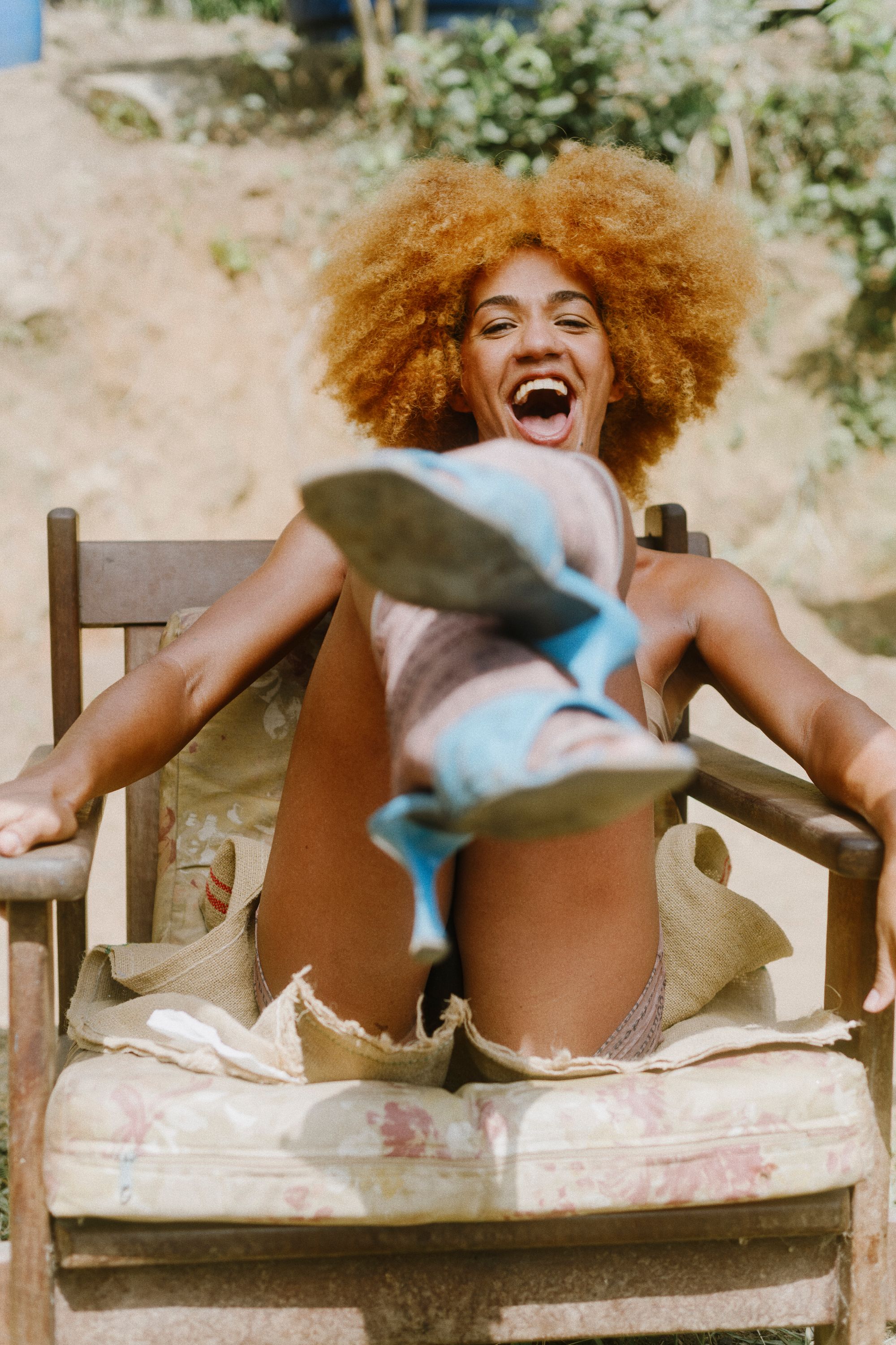 Brasilian travesti with natural red hair, wearing a crystal necklace, is seated on a chair outside in nature, looking at the camera and kicking her foot towards the camera, laughing