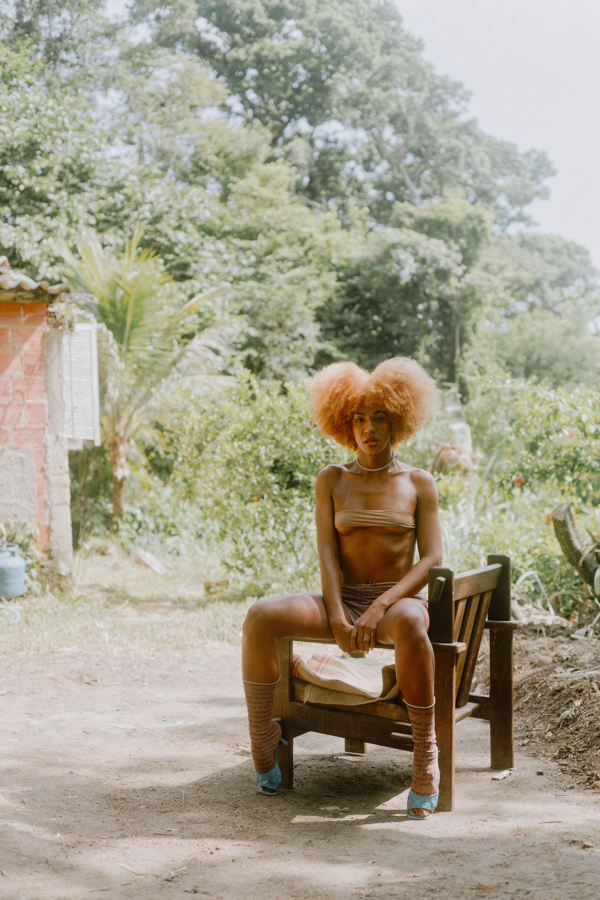 Brasilian travesti with natural red hair, wearing a crystal necklace, is seated on a chair outside in nature, looking at the camera