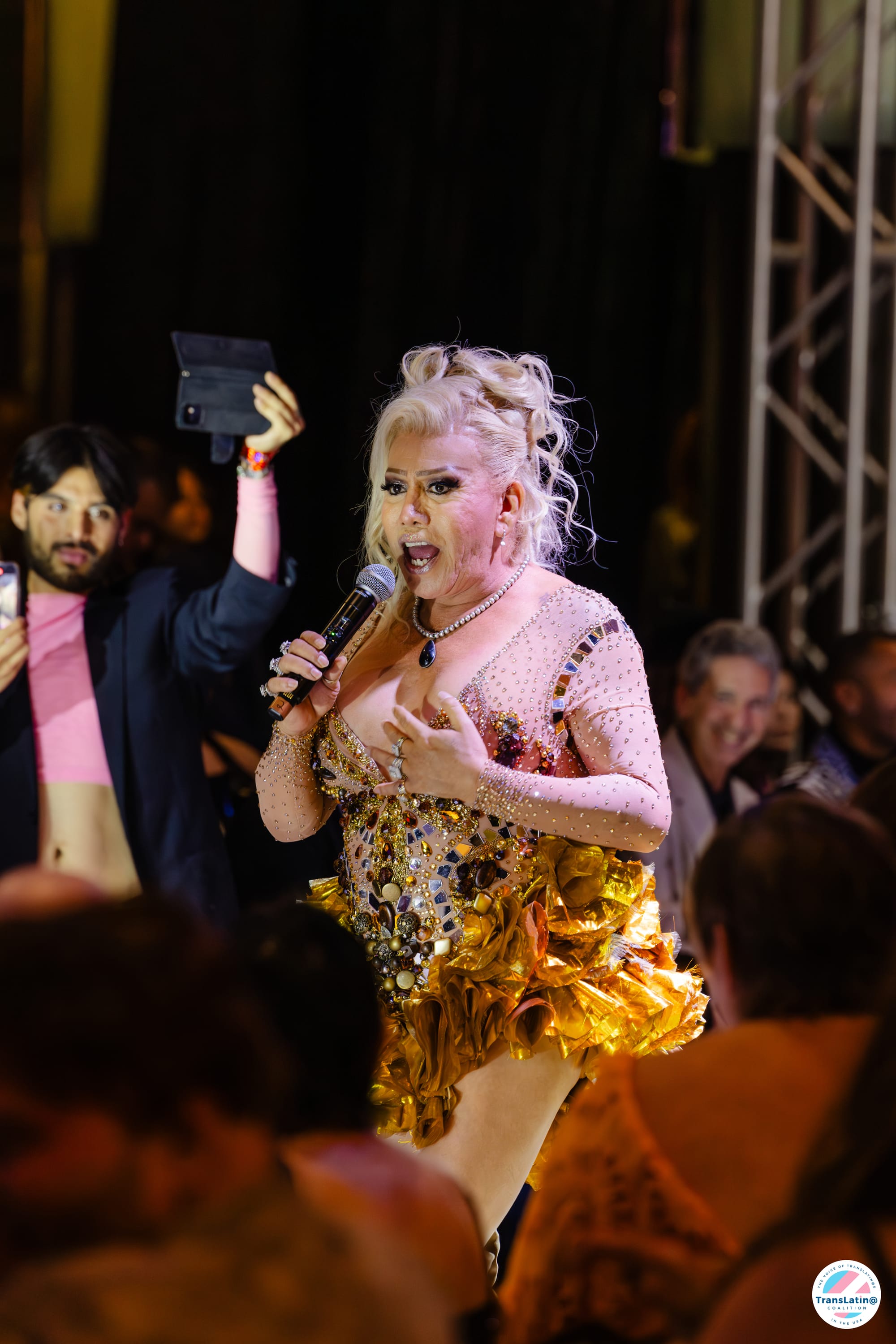 Bamby Salcedo speaking on stage at GARRAS in a jeweled bodice gown with gold metallic ruffles